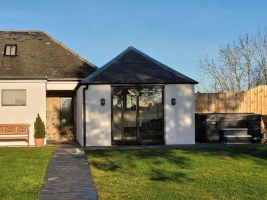 a modern, farm style cottage sits under sunny skies. a hot tub to the right.