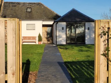Looking up a paved path towards a cottage in a large, sunny garden 